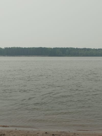 The image depicts a wide river in Bibikha, Russia, under a hazy sky. The foreground shows a sandy shore with small stones and debris. The middle ground is dominated by the rippling surface of the water, which appears greyish-green and somewhat murky. In the background, a dense line of dark green trees forms a forest along the opposite bank of the river. The overall atmosphere is subdued and overcast, suggesting a cloudy or smoky day. There are no people or man-made structures visible in the frame.