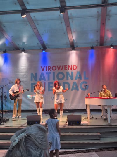This image depicts an outdoor performance taking place on a stage in Differdange, Luxembourg, likely during a festival or event. A banner behind the performers reads "VIROWEND NATIONAL FRIEDA D'DAG". Four individuals are on stage, two women and two men, engaged in a musical performance.
On the left, a man with long blonde hair, dressed in a white shirt and gold pants, plays an acoustic guitar. To his right, a woman with blonde hair, wearing a white mini-dress and white boots, sings into a microphone. Next to her, a woman with red hair, also in a white mini-dress and white boots, sings into a microphone. On the far right, a man in a sparkly gold shirt and white pants plays a white keyboard.
In front of the stage, a young girl in a light blue dress stands facing the performers. In the foreground, the back of a person's head with a white hair clip is visible, obscuring the view of the lower part of the stage and the audience. The stage is lit by various spotlights, with blue and red lighting effects visible. Speakers and microphones are set up on the stage. The overall environment suggests a public gathering.