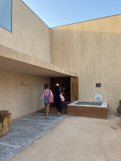 The image captures the exterior of the Museu Praia do Canal, located near Aljezur, Portugal, under a clear blue sky. The building features light tan, smooth stucco walls with a pronounced horizontal overhang above the entrance. Two barefoot individuals, both appearing female, are depicted from behind, walking on a grey stone pathway towards the building's open, dark wooden double doors.

The person on the left wears a pink and white patterned long-sleeved top and a blue denim mini-skirt, carrying a white bag on her left arm. The person on the right wears a black long dress and a white baseball cap, carrying a pink and white patterned bag on her right shoulder.

To the right of the entrance, a rectangular water feature with reddish-brown sides and a light-colored back panel, from which water appears to flow, is situated against the wall. A dark, square plaque is affixed to the wall above this fountain. To the left foreground of the pathway, a partially visible carved wooden object is present. The ground in the foreground is composed of light brown, possibly sandy or gravelly, material. A square window is visible high on the upper left portion of the building's facade.
