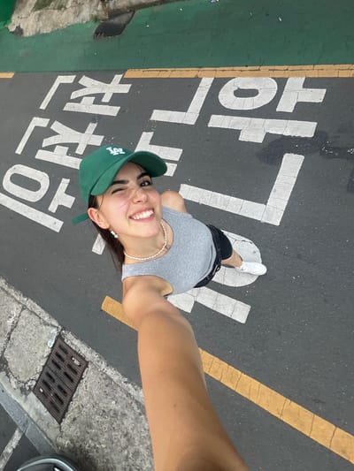 A young woman with dark hair, wearing a grey tank top, black shorts, a green baseball cap, and white sneakers, is posing for a selfie on a street in Daegu, South Korea. She is winking and smiling at the camera, with a pearl necklace around her neck and a pearl earring in her visible ear. The street is marked with white Korean characters, likely indicating a crosswalk or a no-parking zone, and a yellow line runs along the edge of the road. A green painted curb is visible on the upper left of the image, and a metal grate is set into the concrete sidewalk on the lower left. The image is taken from a high angle, looking down at the woman.