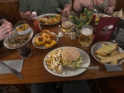 The image shows a group of people having a meal at a restaurant in Broadland, United Kingdom. A wooden table is filled with various dishes, including a burger with fries covered in mayonnaise, onion rings, what appears to be chicken in mushroom sauce with french fries, fish and chips with peas, and cheesy garlic bread. There are also two glasses of beer and a glass of dark liquid. The people are interacting with the food, using forks and knives. A bottle of ketchup, salt and pepper shakers, a small potted plant, and a restaurant menu are also visible on the table. The restaurant appears to be indoors.