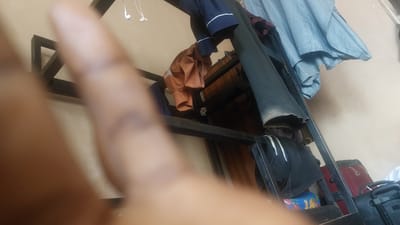 The image depicts an indoor scene, likely a room, with a prominent black metal bunk bed frame. Hanging on the frame are several items of clothing, including what appears to be a light blue, possibly a curtain or bedsheet, and a dark blue garment with white trim. Below these, a rust-colored shirt or jacket is draped over a bar. A black garment with white stripes is also visible hanging on the lower section of the frame. To the right of the bunk bed, a red suitcase and a black bag are stacked. The background is a plain, light-colored wall. In the foreground, a blurred hand is visible, suggesting the image was taken by someone within the scene. The presence of a bunk bed and stacked luggage suggests the location could be a dormitory, guesthouse, or a shared living space. The overall impression is one of a somewhat cluttered, lived-in environment. The location is specified as New Nyanya, Nigeria, a town in the Nasarawa State, which is part of the Abuja Federal Capital Territory region.