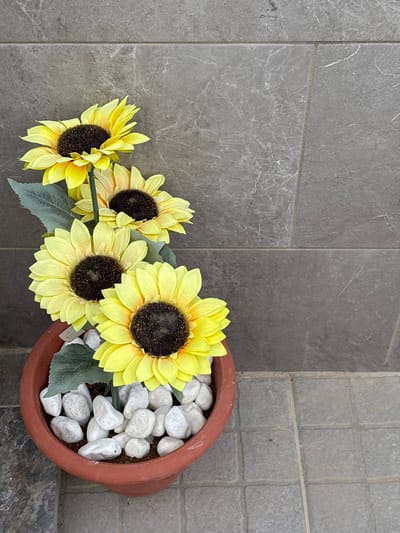 A cluster of four artificial sunflowers in a reddish-brown pot sits on a tiled surface in front of a grey tiled wall. The pot is filled with white pebbles. The sunflowers are arranged vertically, with the largest and most vibrant flower at the bottom. The background consists of large, rectangular grey tiles with a subtle marble-like pattern. The foreground also features grey square tiles. The lighting suggests an outdoor setting, likely during the day, with no shadows indicating direct sunlight. The scene is static and peaceful, with no visible people or animals. The overall impression is one of artificial, decorative floral display.