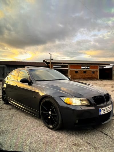 The image shows a black BMW sedan parked on a concrete surface in what appears to be an urban or industrial area. The car's front license plate reads "CB 2223 XP", which is a Bulgarian license plate. The background features a brick building and a sky with clouds and some sunset light. The image is taken at an angle, and the location is likely in Petarch, Bulgaria, as indicated by the license plate.