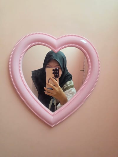 The image shows a young woman wearing a dark hijab and a white shirt with a patterned vest, taking a selfie in a pink heart-shaped mirror. She is holding a smartphone in her right hand, which is reflected in the mirror. The mirror is hung on a plain, light pink wall. The scene appears to be indoors, possibly in a bedroom or a dressing area. No specific location in the City of Medan, Indonesia, can be determined from the image.