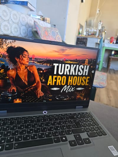 The image shows a laptop on what appears to be a grey couch. The laptop screen displays an image of a woman in a dress in front of a city skyline with the text "TURKISH AFRO HOUSE Mix". The laptop has a grey keyboard with Cyrillic characters in addition to the standard alphabet. The background shows part of a room including a table with items on it, a wall and chair. There is nothing to indicate this photo was taken in Dorkovo, Bulgaria.