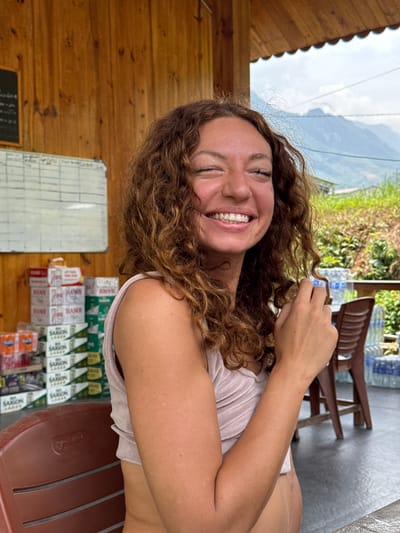 The image, rotated 90 degrees counter-clockwise, features a smiling woman with curly brown hair looking directly at the viewer. She is positioned on her side, with her head towards the left of the frame and her body extending towards the right. She wears a light-colored sleeveless top, and her right arm is bent with her hand near her head. Her expression is bright and happy, revealing white teeth.

Behind her head (to the left in the frame), a wooden wall is visible, along with a white board and a smaller black board, both displaying illegible handwriting. Below her (towards the bottom of the frame), shelves are stocked with numerous packaged goods, including boxes labeled "HANOI" and "SAIGON," likely indicating cigarettes or snacks. A brown plastic chair is partially visible near these items. Above her (towards the top of the frame), the scene transitions to an outdoor view featuring green foliage, distant mountains, and a bright, cloudy sky. Several plastic water bottles and more brown chairs are also seen further back in this area.

The setting appears to be an open-air or semi-enclosed structure, possibly a small shop or guesthouse, situated in a natural, mountainous environment. The visible brands "HANOI" and "SAIGON" confirm the location as Vietnam, specifically Du Già Commune as per context. The bright lighting suggests it is daytime. The woman appears relaxed and is posing for the camera.