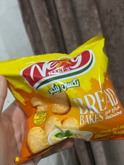 An eye-level, slightly angled shot shows a person holding a vibrant yellow and orange snack package. The package is for "NEXI BAKES," a brand of toasted bread slices, with the Arabic brand name "نكسي بيكس" also visible. The flavor, "Cheese Oregano," is indicated in both English ("Cheese Orega") and Arabic ("جبنة توابل وريحان"). The package design features a prominent red and green "NEXI BAKES" logo on the left side.

On the orange section of the package, various images of small, roundish, golden-brown baked bread pieces (croutons or bread crisps) are displayed. One section shows these bread pieces accompanied by a white dipping sauce, possibly a cheese dip, garnished with green herbs, suggesting the oregano flavor. A small yellow circular emblem near the bottom right of the package contains Arabic text stating "منتج عالي الجودة" (high quality product) and "غير مقلي" (not fried), highlighting a health-conscious aspect of the product.

The person's fingers and part of their palm are visible at the bottom and right side, holding the package. The background is softly blurred, showing horizontal lines, possibly from wooden planks or a textured wall, in muted brown and gray tones. The lighting is soft and even, suggesting an indoor setting, and the focus is clearly on the snack package.