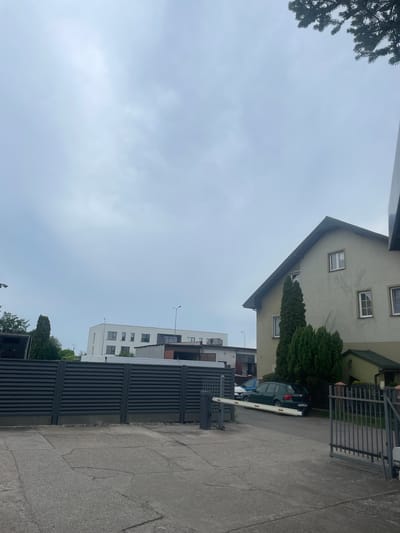 The image shows a street scene in Kołobrzeg, Poland, on an overcast day. In the foreground, a grey, slatted metal fence stretches across the scene, with a concrete driveway in front of it. A white barrier arm, typically used for controlling vehicle access, is lowered across the driveway. To the right of the fence, a grey metal gate is partially open, revealing a glimpse of more fencing and greenery.
Behind the fence, several buildings are visible. On the left, a modern white building with multiple windows stands out. To its right, a light-colored house with a sloping roof and several windows is situated. Trees, including tall, slender evergreens and smaller bushes, are scattered around the buildings.
A dark green car is parked on the driveway, partially obscured by the fence and barrier arm. In the background, other structures and trees can be seen, suggesting an urban or suburban environment. The sky is uniformly grey and cloudy, indicating muted natural light. The overall impression is one of a controlled access area, possibly a residential or commercial property.