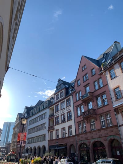 The image depicts a street scene in Frankfurt am Main, Germany, specifically the area near the intersection of Zeil and Goethestrasse (latitude: 50.111, longitude: 8.683). The street is lined with a row of historical buildings with a mix of architectural styles. The buildings are five to six stories high and constructed from brick, stone, and plaster, with large windows and ornate details. The buildings have a reddish-pink and cream color scheme, and they are decorated with decorative iron balconies and railings.
There are several people walking on the sidewalk, some of them in groups, others alone. The sidewalk is lined with trees and street furniture, such as benches and trash cans. In the foreground, a group of people is gathered around a newsstand, likely observing a political campaign poster featuring a man in a suit and tie.
The street is lined with parked cars on both sides, some of them are covered in a layer of dust. A bright blue sky with scattered clouds is visible in the background. The street is illuminated by the sun shining brightly above the buildings.