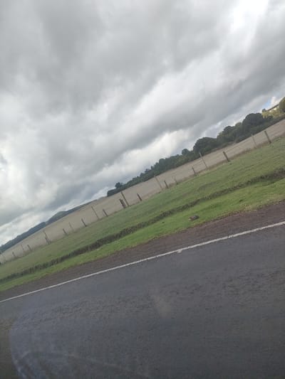 The image shows a rural landscape under a cloudy sky. A dark asphalt road runs diagonally across the foreground, marked with a white line. Beyond the road is a green, sloping field enclosed by a fence made of wooden posts and wire. The field stretches into the distance, rising slightly. Trees line the horizon in the background. The scene suggests a location in a rural area, possibly on the outskirts of a populated region. The provided location of Kisima ward, Kenya, is unlikely to be accurate. There is no immediate evidence of people or animals. The overall impression is of a countryside scene on a cloudy day.