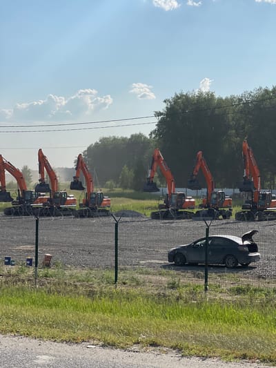The image shows a row of orange excavators parked on a gravel lot. A dark-colored sedan is parked to the right of the excavators, with its driver's side door open and a person's arm visible reaching into the car. The scene is outdoors, with a fence separating the lot from a grassy area. In the background, there are trees and a bright blue sky with scattered white clouds, suggesting it is daytime. The lighting indicates it might be late morning or early afternoon. The excavators appear to be new or well-maintained, all facing in the same direction. The presence of multiple excavators suggests a construction site, equipment rental yard, or similar operational area. The open car door and the arm reaching in imply someone is either getting in or out of the vehicle, or retrieving something from it. No specific text is clearly legible on the excavators or the car. The overall impression is one of a functional, operational outdoor space.