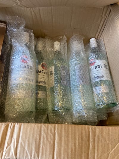 A cardboard box contains five bottles of Bacardi Carta Blanca white rum, each individually wrapped in bubble wrap. The bottles are arranged in a row, with the labels visible on some of them. The box is open at the top, and the corrugated cardboard interior is visible. The scene appears to be a delivery or storage scenario. The location is not specified within the image.