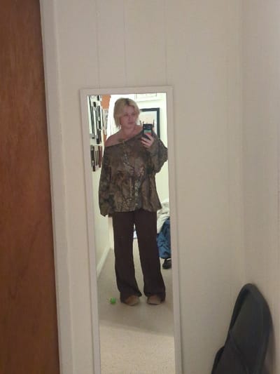 A blonde-haired female is depicted in a full-length mirror selfie, taken in an indoor residential setting within the city of Area D (Elaho/Garibaldi), Canada. She is standing on a light-colored carpet, wearing a loose-fitting, camouflage-patterned, off-the-shoulder long-sleeved shirt, dark brown wide-leg pants, and light brown flat shoes. She holds a dark-cased smartphone in her right hand to capture the image. Visible tattoos are on her left shoulder/chest area. The mirror's reflection shows a wall with multiple framed items to the left and draped blue fabric with a dark shoe to the right. A small green object, resembling a toy frog, rests on the carpet near her feet. Outside the mirror frame, a reddish-brown wooden door or panel is visible on the left, and a light-colored wall with a dark, curved object leaning against it is on the right.