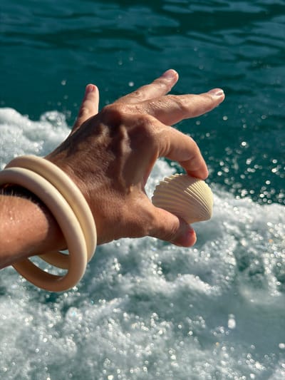 A person's hand, adorned with two cream-colored bangles, holds a small, ribbed seashell against a backdrop of sparkling ocean water. The hand is positioned centrally in the frame, with fingers slightly curved to cradle the shell. The sunlight glints off the water, creating numerous bright, circular highlights that create a bokeh effect. The water itself is a deep turquoise, suggesting it is either the ocean or a large body of water.
The activity appears to be a moment of leisure, possibly on a boat trip, as indicated by the movement of the water and the spray. The bright sunlight and the sparkling water suggest a clear, sunny day, likely during daylight hours. The presence of the seashell and the ocean setting imply a tropical or coastal location. The image conveys a sense of peace and enjoyment of nature. There is no visible text in the image. The location context provided, Baan Thong Krut, Thailand, aligns with these visual cues of a sunny, tropical beach or coastal environment.