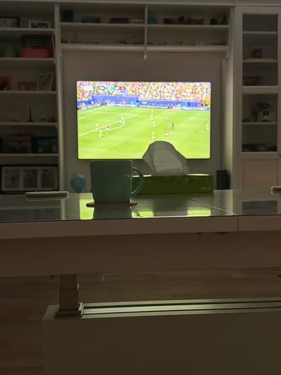 The image depicts a living room setting with a focus on a coffee table and a television screen displaying a soccer match. On the coffee table, a teal-colored mug rests on a coaster, positioned to the left of a green tissue box topped with white tissues. To the left of the mug, another coaster is visible. The television screen, showing a game with players in red and yellow uniforms on a green field, is the dominant background element, framed by white shelving units filled with various items. The overall ambiance suggests a relaxed evening at home. The image does not contain information that can be used to determine its location in Helmond, Netherlands.