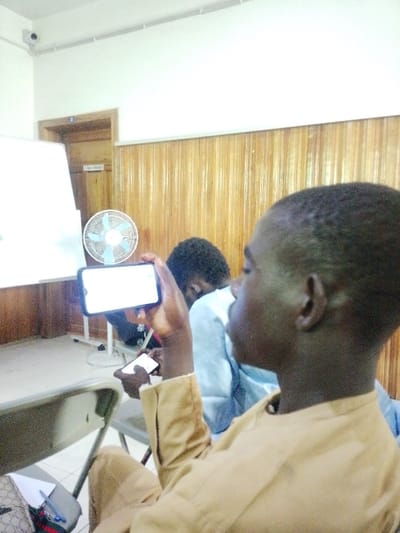 The scene depicts an indoor room, identified as being in Kabuga, Nigeria, consistent with a classroom or meeting space. In the foreground, a dark-skinned male wearing a light brown long-sleeved garment is seated, viewed from his right profile. He holds a black smartphone horizontally with his right hand, displaying a brightly illuminated screen with indistinct white content. His left hand simultaneously holds a smaller, dark-colored smartphone. Behind him and slightly to the left, another individual with dark, curly hair is seated, wearing a light blue top, and appears to be looking downwards.

The room features light brown vertical wooden paneling on the lower portion of the walls, transitioning to an off-white or cream color on the upper walls and ceiling. A white oscillating fan on a stand is positioned in the midground to the left of the foreground subject. Partially visible to the fan's left is a white easel-style whiteboard. A light brown wooden door frame with a closed door, marked with a white number "2", is present in the upper left background. A small, dark security camera is mounted on the off-white ceiling in the upper left corner, with white electrical conduit running along the ceiling. A patterned bag, a pen, and paper are visible on a surface or chair near the foreground subject's feet.