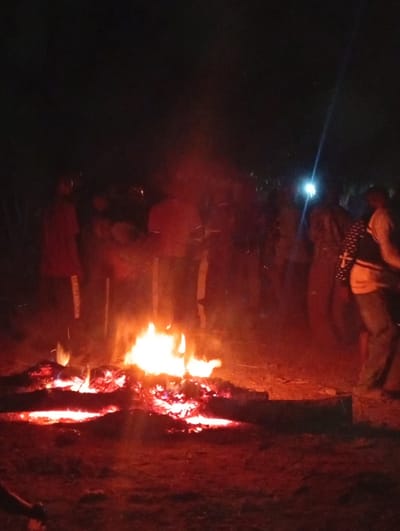 A nighttime gathering around a large bonfire. Several blurred figures stand behind the fire, illuminated by its glow and a bright light in the background. The fire is built with logs and burns intensely, casting a red and orange hue on the surrounding area and the ground. The scene is dimly lit, creating a warm and communal atmosphere.