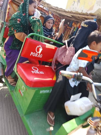 This image depicts multiple individuals riding in a green three-wheeled passenger vehicle, likely an auto-rickshaw, on a sunny day in Shibpur, Bangladesh. The vehicle features red upholstered seats prominently displaying "PORAG" and a stylized flame logo in white text.
On the left side of the vehicle, a woman with a light complexion wears a dark green patterned headscarf over a light brown garment and purple trousers, her left hand gripping the vehicle structure. Adjacent to her, a woman in a black and white striped top and dark head covering holds a smartphone in her left hand while her right hand is raised in a gesture. To her right, a pink backpack is slung over the shoulder of another individual, whose upper body is largely out of frame, but whose legs and feet in brown flip-flops are visible. Towards the front right, a young boy in a light-colored patterned short-sleeved shirt is seated, grasping handlebars that appear to be part of the vehicle's steering or a bicycle. A white plastic container is affixed to the front section of the vehicle.
The background shows a bustling street environment consistent with a market or commercial area, with various goods displayed on stalls or in shops under a makeshift canopy. The ground visible beneath the vehicle appears to be an unpaved or dusty road. The scene is illuminated by strong daylight.
