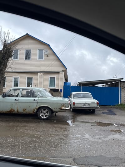 Observed from within a vehicle, the scene depicts a residential area in Ново-Никольское, Russia. Two cars, a light blue sedan with rust and a white sedan with license plate "M630PO 199", are parked on a wet, asphalt road. Adjacent to the road is a two-story, light-colored house with blue trim. To the right of the house, a blue metal building with a gray roof is visible. The sky is overcast, and tree branches are visible in the lower left.