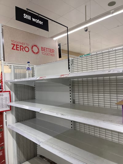 The image shows a supermarket shelf in the "Still water" aisle at a store in Middle Park, Australia. The shelves are empty except for one bottle of water. The shelves are made of metal and have a grid pattern. The sign above the shelf reads "Together to Zero Better Together". The store has a white ceiling with fluorescent lights. The store's security camera is visible in the upper left corner of the image.