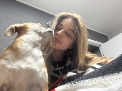 An intimate, low-angle indoor shot captures a young person and a dog resting together. The image is oriented sideways, suggesting it was taken while looking up at them. A person with long, light brown hair and closed eyes appears to be sleeping or deeply relaxed, with their head resting on or under a soft blanket. Their nose has a small piercing. Beside them, a light brown and white dog also has its eyes closed, its head gently touching the person's face, conveying a sense of calm and companionship.
The person is partially covered by a fuzzy blanket with black and white stripes, and a glimpse of red fabric is visible underneath. Under the blanket, the person is wearing a dark garment with stylized white patterns that resemble letters or symbols. The background shows a light-colored ceiling with a molding and a darker wall or section of the room. The lighting appears to be soft and ambient, characteristic of an indoor setting during the daytime. The overall mood is peaceful and serene.
The scene depicts a moment of shared rest or sleep between the person and their pet, likely in a home setting in Ralevo, Bulgaria. No explicit text is clearly legible on the garment, only abstract white shapes.