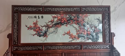 The image shows a wooden framed artwork depicting a large flowering tree with red blossoms, in a room with white textured walls. The art is framed with dark wood with intricate carvings and details in a deep, rich mahogany hue. The artwork is in a room at a location within Hangzhou Shi, China with coordinates 30.449° N, 120.242° E, possibly a home, office, or museum. The image is taken from a frontal perspective, with the artwork occupying the majority of the frame, providing a clear view of the intricate details of the artwork and the surrounding environment. The image is well-lit, highlighting the textures and colors of the scene. There are no people present in the scene. The artwork portrays a large flowering tree with branches reaching out across the canvas, creating a sense of expansive growth. The red blossoms are clustered together in clusters, creating a vibrant contrast against the gray background. The artwork suggests a tranquil and serene setting, evoking a sense of peace and contemplation. The details of the carving on the wooden frame further enhance the sense of craftsmanship and artistry.