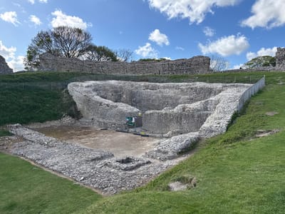 The image captures the extensive stone ruins of a historical structure, likely a castle or fortification, under a clear blue sky dotted with fluffy white clouds. The setting is an outdoor, grassy area, suggesting a historical site or park.
In the foreground and central part of the image, the remains of an excavated or decayed stone structure are visible. This area features low, irregular walls made of light-colored, rough-hewn stones, forming a large, somewhat triangular or semi-circular enclosure. Within this central pit, there are lower, square-shaped stone foundations on a dirt floor. Temporary metal fences, typical of construction or archaeological sites, surround a portion of this central ruin. A small green and white sign is visible behind these fences, partially displaying the text "What's happening?".
Beyond these immediate ruins, higher and more substantial stone walls form a perimeter on an elevated grassy mound. These walls are also constructed from similar irregular stones. Lush green grass covers the slopes leading up to these higher walls and surrounds the entire ruin complex. Several trees, mostly bare-branched with some green foliage, stand behind the distant upper walls, silhouetted against the bright sky. Metal railings, acting as permanent safety barriers, run along the edges of the grassy slopes and around parts of the ruins.
The photograph was taken on a bright, sunny day, likely in the late morning or early afternoon, given the clear light and defined shadows. The overall impression is one of ancient history preserved within a natural landscape, located in the King's Lynn and West Norfolk area of the United Kingdom.