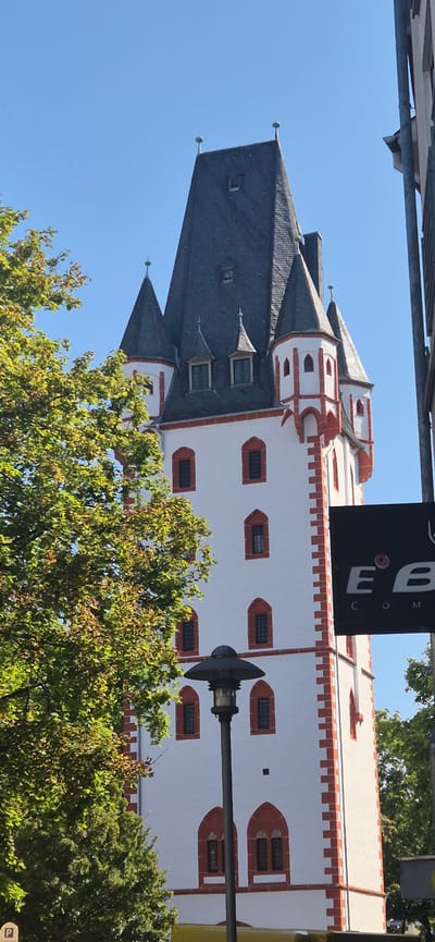An eye-level, slightly low-angle, full shot shows the white, historic whitewashed tower of the Hof zum St. Peter, also known as the Mainzer Stadtschloss, located in Mainz, Germany. The tower is a white structure with red-trimmed windows and architectural details. It has a tall, dark grey, pointed roof with several smaller conical turrets. In the foreground to the left, a large green tree with some autumnal color fills a significant portion of the frame. To the right, a black sign with white lettering that reads "EB COM" is partially visible. A black lamppost stands in front of the tower, and the sky is a clear, bright blue. The image captures a sunny day in a European city.