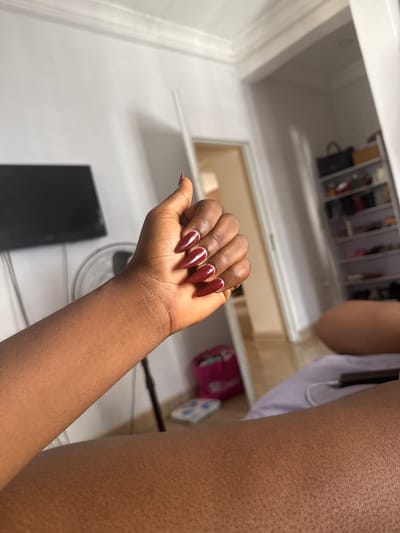 A close-up, indoor shot focuses on a dark-skinned person's hand with long, stiletto-shaped, burgundy nails. One nail has a white stripe detail. The hand and arm are prominent, extending from the lower right corner towards the center of the image. The background reveals a white interior with a partially opened cabinet containing miscellaneous items. Beneath the cabinet is a white wall with a black, rectangular object and a white fan. The overall setting appears to be a room or closet. There is no evidence to support a location in Dushepe, Nigeria.