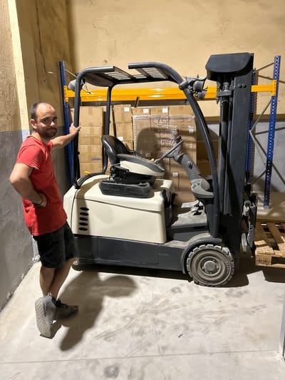 A bald man wearing a red t-shirt and dark shorts stands in a warehouse, leaning on the frame of a forklift. He has his left hand on the forklift's uprights and his right hand on his hip. The forklift is beige and gray, with a black overhead guard. Behind the forklift are shelves stacked with cardboard boxes. The floor is concrete and shows tire marks. The walls appear to be painted concrete. The overall lighting suggests an indoor industrial setting.