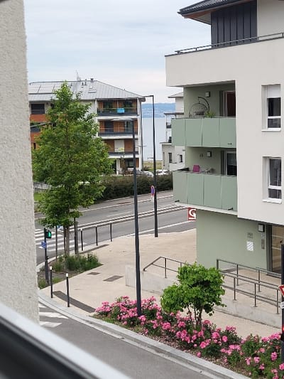 The scene shows a street corner in Publier, France, at coordinates 46.395, 6.54. A street intersection with a traffic light can be seen in the foreground. A sidewalk area with a railing and a small tree leads to a modern apartment building with green and white siding. The view from the window shows another apartment building across the street. The apartment building on the left side is made of red brick and has a balcony. The view from the window also reveals a body of water in the distance, suggesting the location is near a lake or river. The sky is partly cloudy. The image appears to be taken from an upper-level window facing the street corner.
