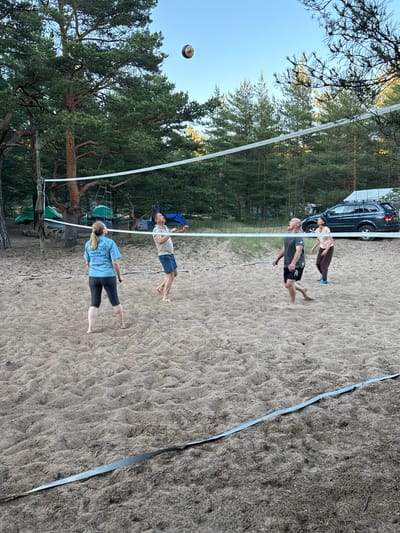 A group of four individuals are playing volleyball on a sandy court in Ozerki (Krasnodolinskaya), Russia. The court is set up with a net, and the players are in various stages of hitting or observing the ball. Two men and two women are present. One woman in a blue t-shirt is positioned on the left side of the net, facing away from the camera. A man in a light-colored polo shirt and blue shorts is on the left side, looking up at the ball. On the right side of the net, a man in a dark t-shirt and black shorts is in mid-action, appearing to hit the ball. Behind him, a woman in a pink t-shirt and brown pants is also looking up, presumably at the ball. The background features a line of pine trees and a dark-colored SUV parked behind the court. Tents and camping equipment are visible further in the background, suggesting a recreational or camping area. The sky is clear and blue.