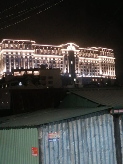 The image shows a grand, multi-story building illuminated by many lights at night. The building has a symmetrical facade with columns and numerous windows. In the foreground, there are structures made of corrugated metal, which appear to be sheds or temporary buildings. One of these structures has two signs attached to it, one with red text and another with blue text displaying a phone number. The sky is dark, indicating nighttime. The location is identified as Lam Vien - Da Lat, Vietnam.