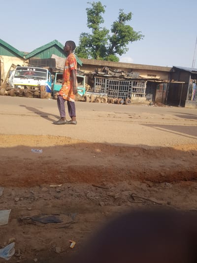 A young man in a patterned orange shirt and dark pants walks along a dirt and asphalt road in Kufena, Nigeria. He is facing to the right and casting a shadow to his left. In the background, there are several disassembled truck chassis and truck parts arranged in front of a low-rise building with a green corrugated roof. The sky is clear and blue. The foreground shows a dirt embankment.