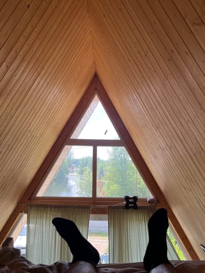 The image shows a triangular window in a wooden-paneled room, with a view of a body of water and trees outside. The perspective is from a bed, with two feet wearing black socks visible in the foreground. There are light green curtains covering a portion of the lower window. A black TV mount is visible on the wall above the curtains. The scene is likely a bedroom in a house or cabin located in or near Vasilyevo, Russia, given the context.