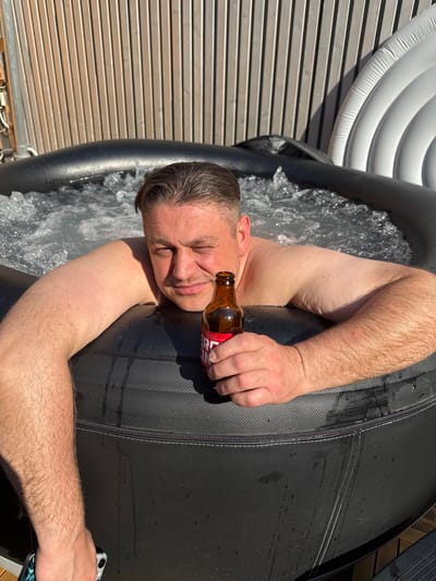 The media file shows a man relaxing in an outdoor hot tub. He is holding a brown glass beer bottle with a red and white label. The man is leaning back with his eyes closed, suggesting a relaxed or contented emotional state. Water is splashing around him, indicating the hot tub is in use. The setting appears to be a backyard or patio area, with a wooden fence visible in the background and the inflatable structure of the hot tub taking up a large portion of the frame. The lighting suggests it is daytime, likely sunny. Visible text on the beer bottle's label includes "APERITIVO". The location context provided is Bevern, Germany.