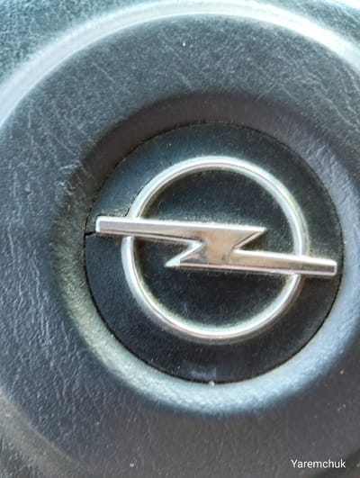 This media file is a close-up, eye-level shot of an Opel car emblem on a dark, textured surface, likely a steering wheel or dashboard. The emblem, a silver lightning bolt within a circle, is the primary subject. The surrounding surface appears to be worn, with visible scratches and dust, suggesting it's part of a used vehicle. The lighting is bright, casting subtle highlights on the metallic emblem and the textured surface. There are no people or other objects visible in the frame. The text "Yaremchuk" is present in the bottom right corner, appearing to be a watermark or signature. Given the provided location context of Zhytomyr, Ukraine, the scene is likely within a car in that region. No specific activity or event is depicted, it's a static image of a car's interior detail. The time of day and weather are not discernible from the image.