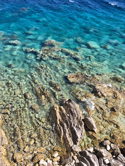 The image shows a close-up, high-angle view of a rocky shoreline meeting clear, turquoise water. The water is shallow enough to reveal the underwater rocks and seabed, which are a mix of light and dark stones. Small waves are visible, gently lapping at the shore. The rocks on the shore are varied in size and texture, with some appearing smooth and others rough and jagged. There are no people or animals visible in the scene. The environment is natural and appears to be a coastal area. The scene is consistent with the coastline of Perazića Do, Montenegro, known for its clear waters and rocky shores.