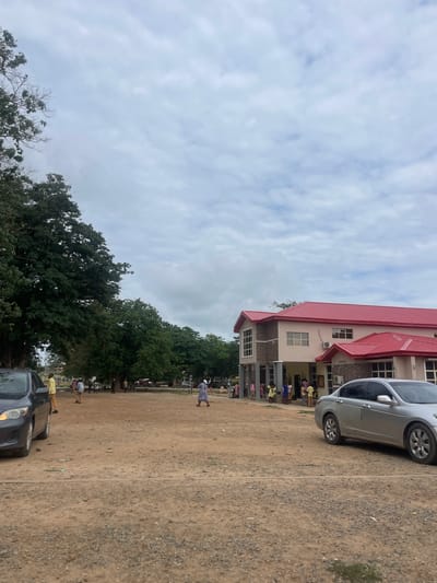 This image depicts an outdoor scene in Karu, Nigeria, characterized by an unpaved, light brown dirt and gravel ground surface with scattered small patches of green vegetation. The sky is overcast with light grey clouds.
On the right side of the frame, a two-story building with a prominent bright red corrugated metal roof is visible. The building's walls are light beige, with sections of grey stone cladding on the lower level and around window frames. Multiple rectangular windows are present, and a front entrance area features structural columns and an overhang. Two external air conditioning units are mounted on the building's facade.
To the left, a dense line of large, leafy green trees extends from the midground into the background, with some bare branches visible in the upper left corner.
Two sedans are present: a dark grey sedan is partially visible on the far left, facing towards the right. A silver sedan is parked on the right side, facing towards the left, with its side profile prominent.
Approximately 15-20 individuals are dispersed across the scene. A group of people, including adults and several younger individuals, stands near the front entrance of the building on the right. Other individuals are seen walking across the open dirt area; for example, one person in a yellow top and dark trousers stands near the dark grey car on the left, and another person in a light grey tunic and white head covering walks towards the left on the right side of the frame. Clothing visible includes tunics, trousers, and dresses in various colors.