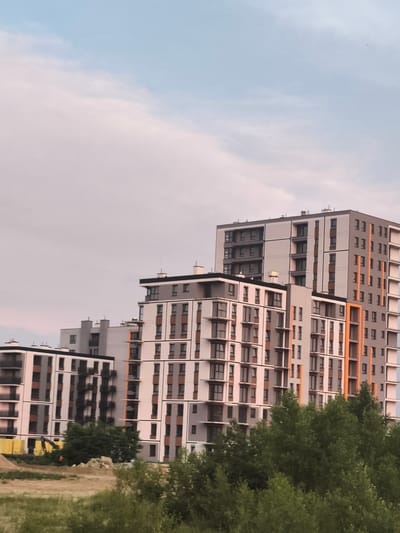 This is a photograph of a modern apartment complex in Nowy Dwór Mazowiecki, Poland. The buildings are tall and multi-story, featuring a geometric design with alternating white, grey, and brown facades. Many windows and balconies are visible, some with dark frames and others with lighter ones. Vertical orange accents add a pop of color to one of the buildings. In the foreground, lush green trees and vegetation partially obscure the lower levels of the structures. The sky above is a pale blue with scattered clouds, suggesting it is daytime. Some construction materials, including yellow tarps and a pile of dirt, are visible on the ground, indicating ongoing development in the area.