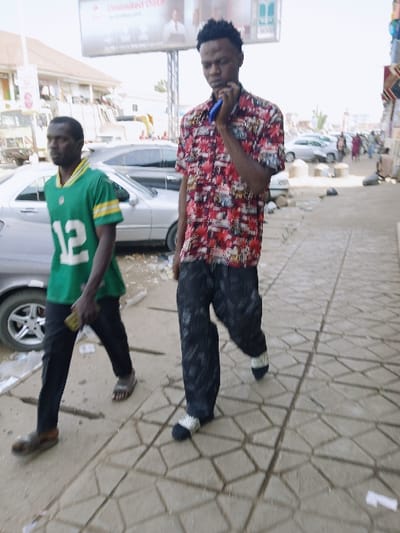 The image depicts two adult males walking on a geometrically patterned concrete sidewalk in an urban environment during daylight.
The taller male, positioned in the foreground and towards the right, is wearing a short-sleeved, collared shirt patterned in red, black, and white abstract motifs, dark patterned trousers, and white socks with dark tips, without visible shoes. His right hand holds a blue object, possibly a mobile phone, near his chin, suggesting engagement with the device. He is looking downwards.
The second male, positioned to the left and slightly behind the first, is wearing a green American football jersey with a yellow collar and yellow stripes on the sleeves, featuring a white number "12" on the front. He wears dark trousers and dark, open-toed sandals. He holds a small, rectangular object in his right hand and looks straight ahead.
In the background, multiple vehicles, including silver sedans, are parked on a street or lot adjacent to the sidewalk. Multi-story buildings, some with visible windows and balconies, line the background. A large billboard or sign displaying "Unlimited Data" with an image of a person is visible above the buildings. The sidewalk shows some scattered debris. There is no visual information present in the image to identify the location as Kungulu School, Zambia; the scene appears to be a general city street.