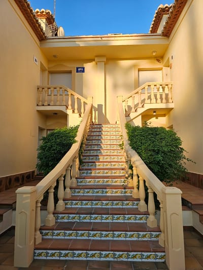 The image shows an external staircase leading to two identical doorways. The staircase has a cream-colored balustrade and is decorated with colorful tiles featuring a yellow, green, and blue ornate pattern. Lush green bushes are planted on either side of the staircase. The building walls are painted a light yellow, and the roof has terracotta tiles. A blue sign with "IP" in white is visible on the left side of the building. The scene is illuminated by sunlight, casting shadows on the stairs and walls. This is likely a residential building or apartment complex in Deveses, Spain.