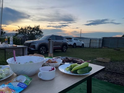 A group of people are having a barbecue dinner in the city of Yermekeyevo, Russia. The sky is a mix of blue and orange, indicating that the sun is setting. The table is laden with food and drinks. There are several cars parked in the background.