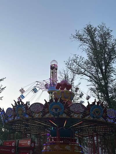 This is a photograph of an amusement park ride in Manas, Kyrgyzstan. The ride appears to be a carousel-style swing ride, with colorful lights and decorative elements. In the background, a tall tower with lights is visible, as well as several swings in motion with people riding them. The sky is clear and blue, suggesting it is daytime. Trees are visible in the background, indicating an outdoor setting. Signs with Russian text are present in the foreground, with one reading "МАЛЫШКОВЫЙ ЗОНА" which translates to "KIDS ZONE." The overall scene depicts a lively and festive atmosphere characteristic of an amusement park.
