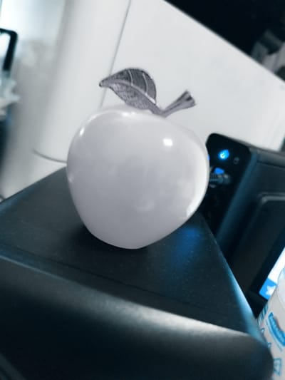 The image shows a close-up view of a white, glossy apple-shaped decorative object with a silver leaf on top, placed on a dark surface. To the right of the apple, a black electronic device is visible, with a blue LED light illuminated on its surface. The background is blurred and appears to be an indoor environment with white and grey elements, possibly furniture or walls. The overall lighting suggests it is daytime. The scene is devoid of any people or recognizable landmarks of Skadovsk, Ukraine, or any other city.