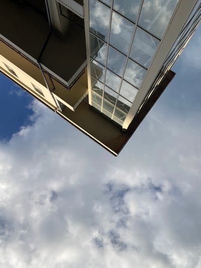 The image is an upward-angle, low-light photograph of a modern building exterior against a cloudy sky, taken in Гаспринский поселковый совет, Ukraine. The building features a light-colored facade with several small, rectangular windows. A prominent section of the building is clad in glass panes, reflecting the sky and clouds. The lower portion of the building has a darker, ribbed siding. A gutter system is visible on the side of the building. The sky is filled with a mix of dark and light gray clouds, with patches of blue sky visible. The orientation of the photograph suggests it was taken looking directly up at the building. There are no people or visible signs of activity.