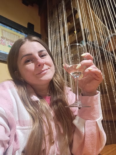 A woman with long brown hair and a pink and white jacket is holding a wine glass with liquid in it. The background includes a picture frame with artwork and beaded curtains. It's plausible this image was taken in Chernogolovka, Russia, if the woman was located there and the setting is consistent with establishments or residences in that city, though the image itself doesn't explicitly confirm the location.