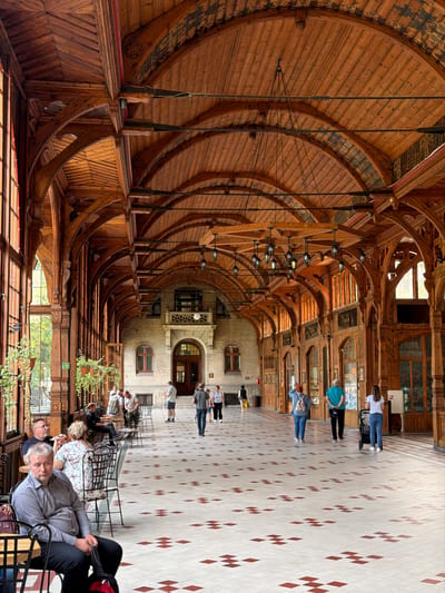 The image shows the wooden colonnade of the Health Resort in Świeradów-Zdrój, Poland. The colonnade features elaborate wooden architecture with arched ceilings, intricate carvings, and hanging lamps. The floor is tiled with a pattern of white and red squares. Several people are present in the scene, including individuals sitting at tables, walking, and pushing a stroller. On the left side, a man is seated at a table, facing the camera, with a pensive expression. Further down, other patrons are seated at tables, some eating or conversing. In the center and right of the image, people are walking along the colonnade, some from behind. The architecture suggests an older, possibly historic building, with large windows offering glimpses of greenery outside. The atmosphere appears relaxed, with people enjoying the covered walkway and the surrounding environment.
