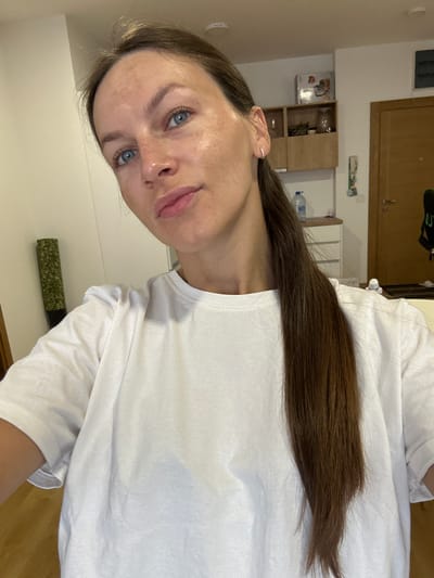 A medium shot captured from a slightly elevated angle shows a woman with long, dark brown hair pulled back into a ponytail. Her blue eyes and fair skin with freckles are visible. She is wearing a plain white t-shirt. The background appears to be an indoor residential setting, with a wall, shelving unit, and a doorway visible. The overall lighting suggests an indoor environment. The image is consistent with a personal photograph taken within a domestic space in Podostrog, Montenegro.